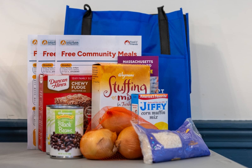 A variety of thanksgiving food items in front of a reusable grocery bag, including beans,stuffing, cornbread mix, and rice, next to a stack of that flyers say 